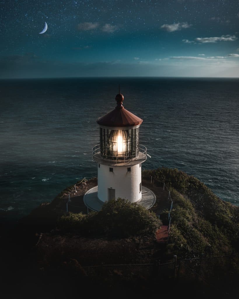 A beautiful aerial shot of a lit lighthouse on a green hill with the half-moon in the beautiful night sky and the ocean in the background